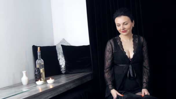 Frumoasă femeie tânără cu păr lung și într-un peignoir negru și decolteu se uită la camera de lângă pahare de vin și o sticlă — Videoclip de stoc