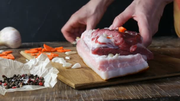 Las manos hembras apuñalan con un cuchillo y condimentos rellenos con rodajas de ajo y zanahorias un trozo de carne fresca cruda de cerdo o tocino en un estilo rústico en tablero de cocina de madera. — Vídeos de Stock