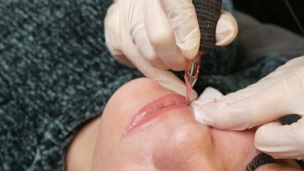 Microblading lip tattoo dengan pigmen pewarnaan khusus yang mengoreksi warna bibir di klinik kosmetologi. Tangan membuat prosedur makeup bibir permanen menerapkan riasan pigmen di bibir dengan mesin tato — Stok Video
