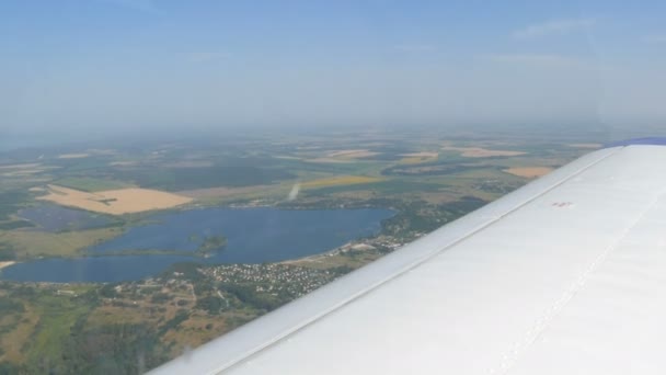 Η λευκή πτέρυγα ενός επιβατικού αεροπλάνου με θέα την κορυφή της φύσης φόντο μιας όμορφης γαλάζιας λίμνης, πετά πάνω από μια λίμνη, πράσινα δάση και μικρά σπίτια και πόλεις. Ταξιδιωτική έννοια — Αρχείο Βίντεο