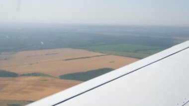 Küçük bir yolcu uçağının penceresinden beyaz bir kanadın arka planına bakın. Sarı tarlalar, yeşil ormanlar