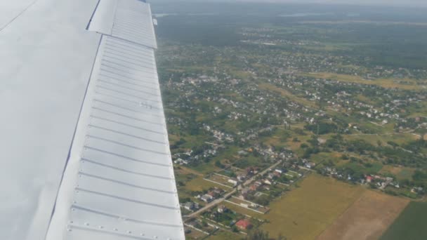 Η λευκή πτέρυγα ενός επιβατικού αεροπλάνου με θέα την κορυφή της φύσης φόντο μιας όμορφης γαλάζιας λίμνης, πετά πάνω από μια λίμνη, πράσινα δάση και μικρά σπίτια και πόλεις. Ταξιδιωτική έννοια — Αρχείο Βίντεο