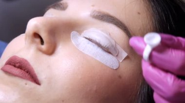 Profesyonel bir güzellik salonunda kirpik temizleme işlemi yapan genç bir kızın yüzü. Usta kıvırma işlemi sırasında kirpikleri özel bir aynayla kontrol eder.