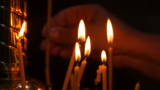 Velas largas y delgadas de cera arden con una llama en una iglesia ortodoxa, rituales conmemorativos para los cristianos. la mano de las mujeres pone una vela en un candelero especial — Vídeo de stock