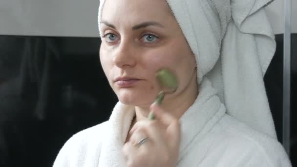 Piękna kobieta z ręcznikiem na głowie robi masaż jadeitowym wałkiem do twarzy z kamienia w łazience. Chińskie narzędzia piękności. Masaż limfatyczny skóry przed starzeniem się zmarszczek — Wideo stockowe