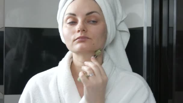 漂亮的女人头戴毛巾，在浴室里用玉石滚筒按摩脸蛋。中国的美容工具。淋巴引流皮肤按摩预防皱纹老化 — 图库视频影像