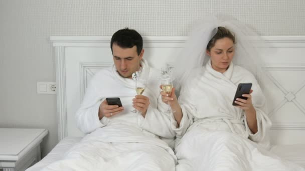 Um jovem casal de recém-casados na cama olha para smartphones batendo copos de champanhe sem olhar um para o outro. Uma noiva num véu, um casal de roupões brancos. Problema da comunicação — Vídeo de Stock