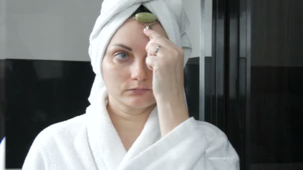 漂亮的女人头戴毛巾，在浴室里用玉石滚筒按摩脸。中国的美容工具。淋巴引流皮肤按摩预防皱纹老化 — 图库视频影像