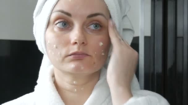 Молода красива жінка наносить крем на обличчя у ванній кімнаті, з білим рушником на голову і в білий халат. Догляд за шкірою обличчя, профілактика раннього старіння та зморшок — стокове відео