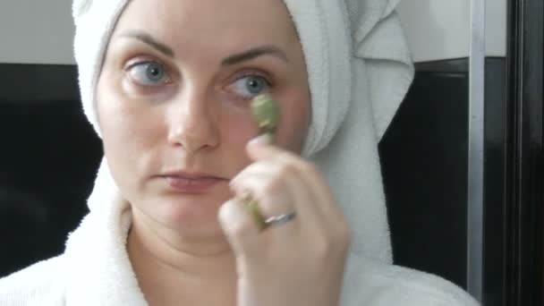 Гарна жінка з рушником на голові масаж з нефритовим роликом на обличчі природного зеленого каменю у ванній кімнаті. Китайські інструменти краси. Лімфатичний масаж шкіри від старіння зморшок. — стокове відео