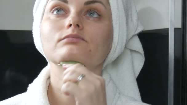 Красивая женщина с полотенцем на голове делает массаж нефритовым роликом для лица натурального зеленого камня в ванной комнате. Китайские косметические инструменты. Лимфатический дренажный массаж кожи от старения морщин — стоковое видео