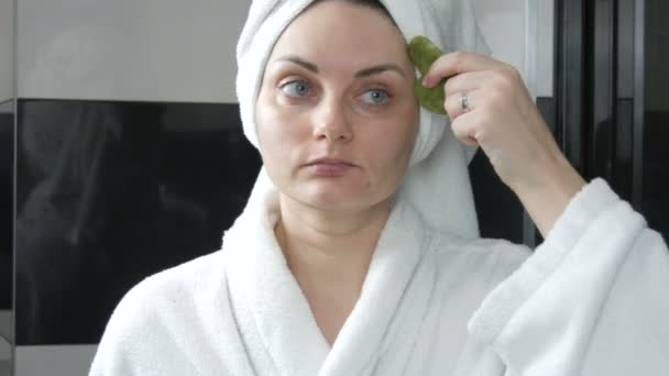 漂亮的女人，头戴毛巾，用玉石刮胡刀按摩，在浴室按摩古奇额头脸。中国的美容工具。淋巴引流皮肤按摩预防皱纹老化 — 图库视频影像