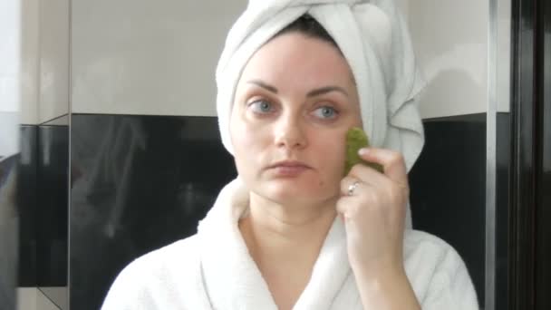 Mooie vrouw met handdoek op het hoofd doen massage met een jade roller voor het gezicht van een natuurlijke groene steen in de badkamer. Chinees schoonheidsgereedschap. Lymfatische drainage huidmassage tegen verouderende rimpels — Stockvideo