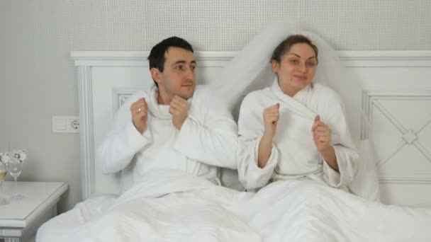 Engraçado casal jovem recém-casados em um hotel alegremente dançando na cama. Noiva em um véu e um roupão de banho branco com o noivo se divertindo e se alegrando — Vídeo de Stock