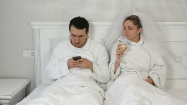 Pareja joven casada divertida de recién casados en el hotel se acuestan en la cama. El novio juega en el smartphone y la mujer en el velo bebe de las copas de boda, exigiendo la atención y celoso del teléfono — Vídeos de Stock