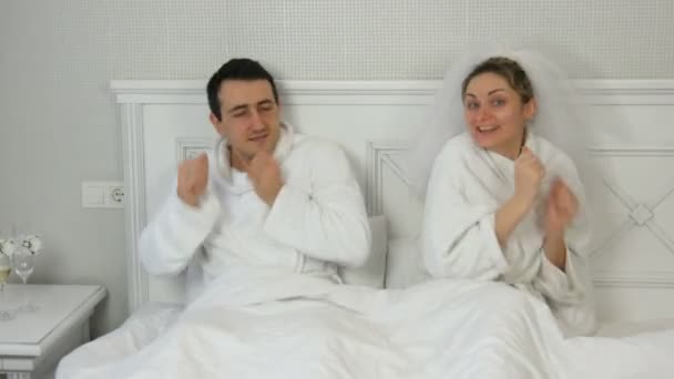 23 de enero de 2021 - Kamenskoe, Ucrania: Pareja joven casada divertida recién casada en un hotel bailando alegremente en la cama. Novia con velo y albornoz blanco con el novio divirtiéndose y regocijándose — Vídeos de Stock