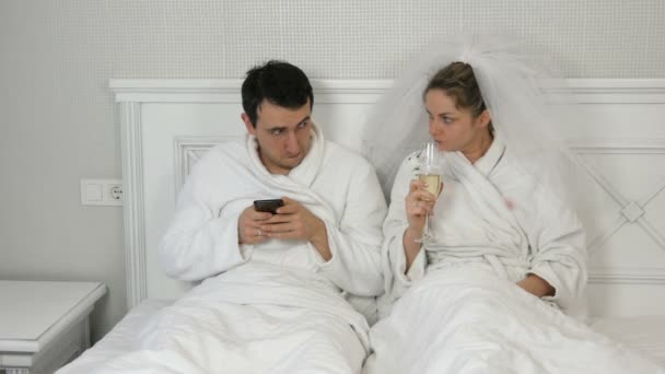 Смішно одружена молода пара молодих в готелі лежить на ліжку. Наречений грає на смартфоні і дружина в куртці п'є з весільних окулярів, вимагаючи уваги і ревності до телефону — стокове відео