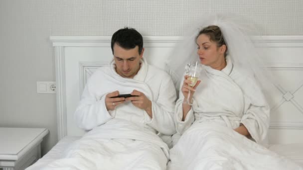Pareja joven casada divertida de recién casados en el hotel se acuestan en la cama. El novio juega en el smartphone y la mujer en el velo bebe de las copas de boda, exigiendo la atención y celoso del teléfono — Vídeos de Stock