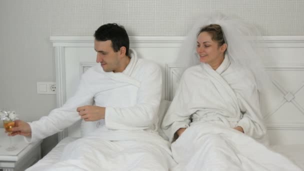 Забавная супружеская пара молодоженов в отеле лежат на кровати и пьют шампанское из свадебных очков. Невеста в вуали и белый халат с женихом веселятся и радуются поцелую — стоковое видео