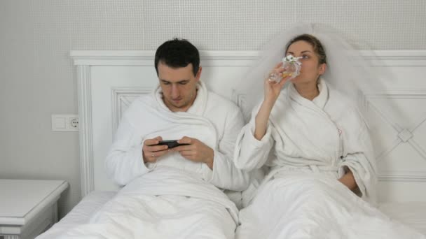 Pareja joven casada divertida de recién casados en el hotel se acuestan en la cama. El novio juega en el smartphone y la mujer en el velo bebe de las copas de boda, exigiendo la atención y celoso del teléfono — Vídeos de Stock