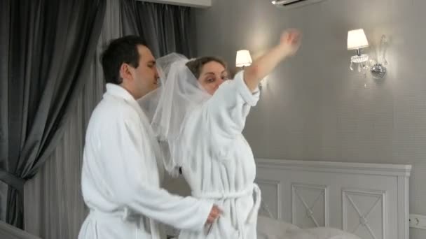 Funny married young newlywed couple dancing merrily in the hotel. Bride in a veil and a white bathrobe with the groom having fun and rejoicing — Stock Video