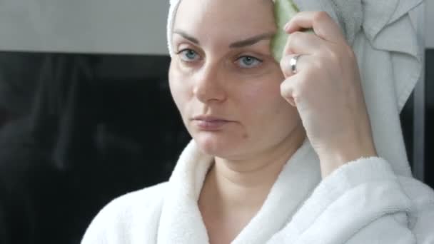 Красивая женщина с полотенцем на голове делает массаж нефритовым каменным скрапером для лица Гуашь Массаж в ванной комнате. Китайские косметические инструменты. Лимфатический дренажный массаж кожи от старения морщин — стоковое видео