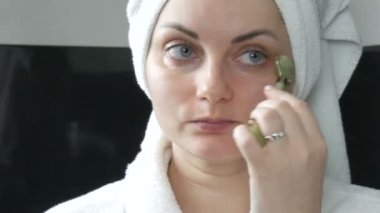 Kafasında havlu olan güzel bir kadın banyodaki doğal yeşil taşın yüzüne yeşim taşıyla masaj yapıyor. Çin güzellik aletleri. Yaşlanan kırışıklıklara karşı lenfatik deri masajı