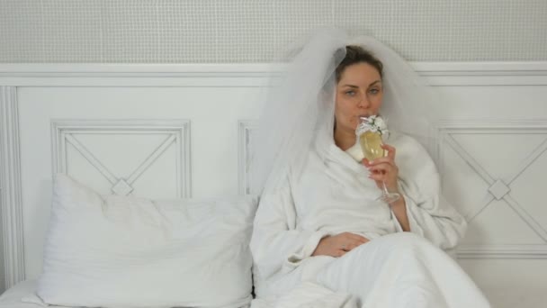Rolig ung brud i en slöja på en säng i ett hotell drycker från bröllopsglas, tittar frestande på kameran och ler — Stockvideo