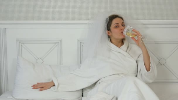 Rolig ung brud i en slöja på sängen på hotellet drycker från bröllopsglas, frestande smeka kudden och vinkar till henne — Stockvideo