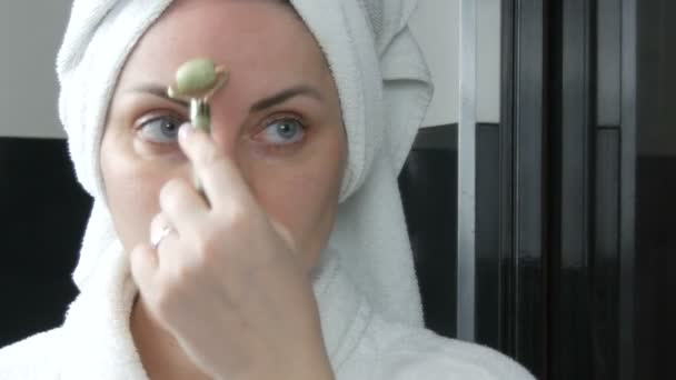 Mooie vrouw met handdoek op het hoofd doen massage met een jade roller voor het gezicht van een natuurlijke groene steen in de badkamer. Chinees schoonheidsgereedschap. Lymfatische drainage huidmassage tegen verouderende rimpels — Stockvideo