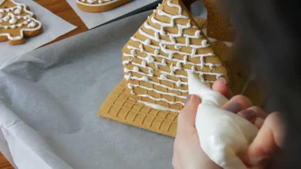 Kvinna dekorerar pepparkakshus med bakverk väska vitt socker söt glasyr, händer på vit tegel bakgrund. Matlagning, bakning hembakat pepparkakshus för jul semester. Traditioner för det nya året — Stockvideo