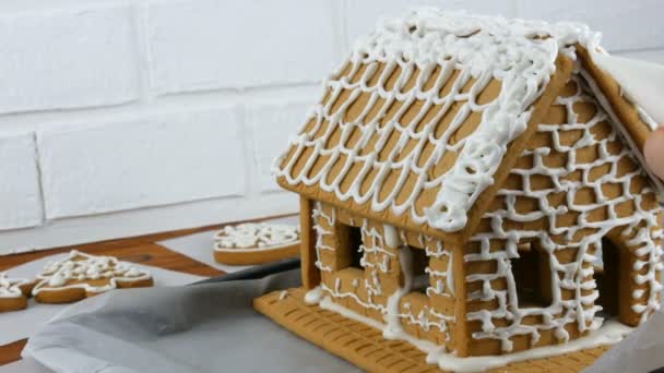 Mulher decora casa de telhado de biscoito de gengibre com saco de pastelaria glacê de açúcar branco, mãos no fundo de tijolo branco. Cozinhar, assar caseiro casa de gengibre feriados de Natal. Tradições de Ano Novo — Vídeo de Stock