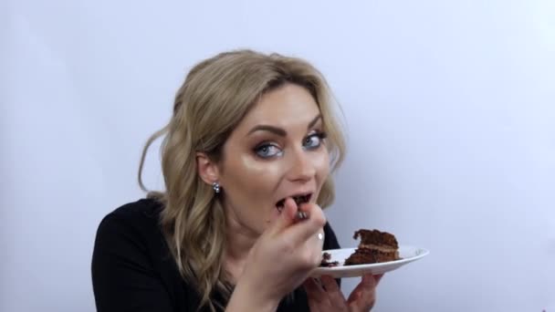 Mooie jonge vrouw model grappig eten brownie chocolade taart op witte achtergrond. jong meisje met mooi smoky oog make-up en zwart avond jurk — Stockvideo