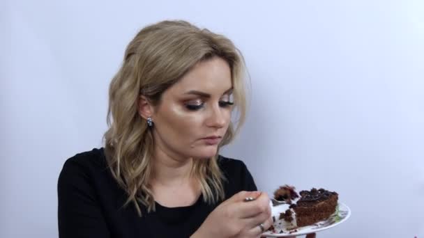 Mooie jonge vrouw model grappig eten brownie chocolade taart op witte achtergrond. jong meisje met mooi smoky oog make-up en zwart avond jurk — Stockvideo