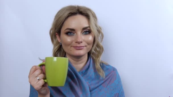 Krásná mladá žena model drží zelený šálek čaje nebo kávy v rukou, pije to, těší a zamyšleně se dívá na bílém pozadí. Mladá dívka s krásným kouřem oční make-up a modrá šála na ramenou — Stock video