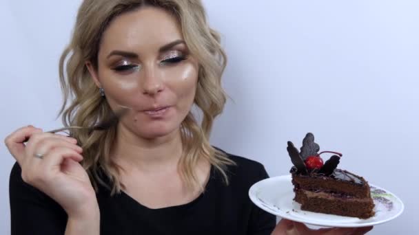 Belle jeune femme modèle drôle manger brownie gâteau au chocolat sur fond blanc. Jeune fille avec beau maquillage des yeux fumé et robe de soirée noire. Apprécie et regarde la caméra — Video