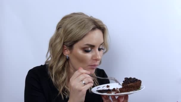 Mooie jonge vrouw model grappig eten brownie chocolade taart op witte achtergrond. Jong meisje met mooie rokerige oogmake-up en zwarte avondjurk. Snelheidsvideo — Stockvideo