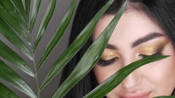 Modelo jovem muito bonita olha através de folhas de palma verde posando no estúdio de fotografia. Retrato de uma menina com uma maquiagem olho fumegante noite brilhante, cabelos escuros e olhos castanhos olha — Vídeo de Stock