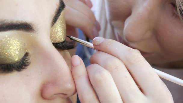 Menina estilista make-up make-up artista aplica uma linha fina de delineador preto em maquiagem brilhante em tons de ouro no olho modelos, close-up view — Vídeo de Stock