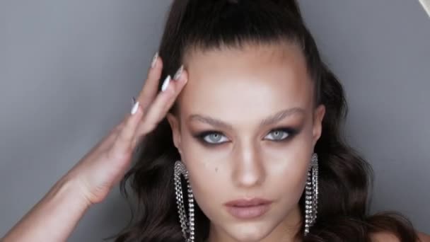 Beau modèle jeune fille avec une coiffure à queue de cheval haute et des boucles d'oreilles massives en strass. Poser à haute couture avec soirée élégante maquiller les yeux fumés. Extension de cils longs et maquillage des yeux — Video