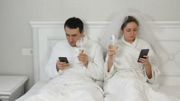 Een jong stel pasgetrouwden op het bed kijkt naar smartphones met glazen champagne zonder elkaar aan te kijken. Een bruid in een sluier, een paar in witte badjassen. Communicatieprobleem — Stockvideo