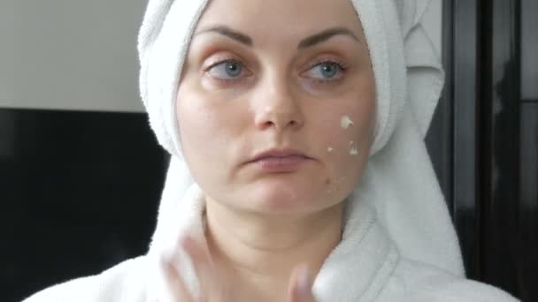 Молодая красивая женщина наносит крем на лицо в ванной комнате, с белым полотенцем на голове и в белом халате. Уход за кожей лица, профилактика раннего старения и морщин — стоковое видео