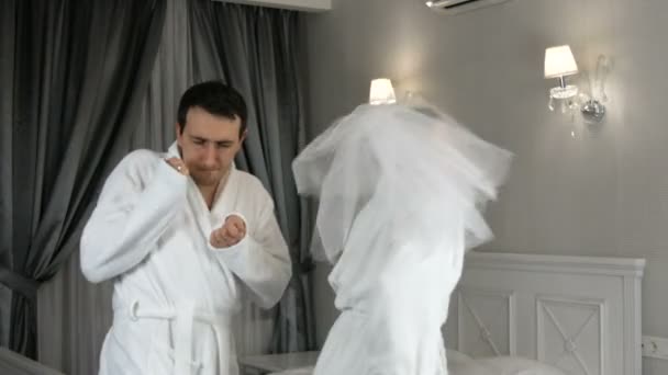 Grappig getrouwd jong pasgetrouwd stel dat vrolijk danst in het hotel. Bruid in een sluier en een witte badjas met de bruidegom plezier en vreugde — Stockvideo