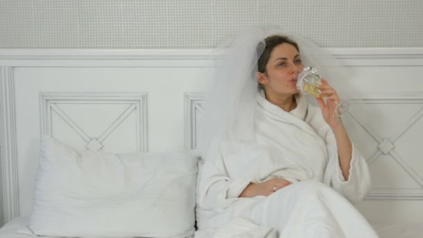 Забавная молодая невеста в вуали на кровати в отеле пьет из свадебных очков, соблазнительно гладит подушку и манит ее — стоковое видео