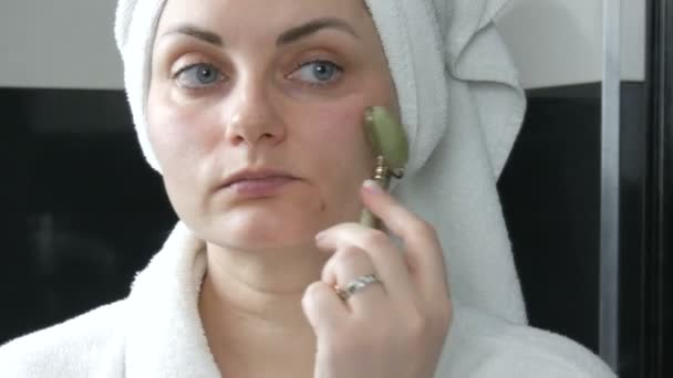 Hermosa mujer con toalla en la cabeza haciendo masaje con un rodillo de jade para la cara de una piedra verde natural en el baño. Herramientas de belleza chinas. Drenaje linfático masaje de la piel contra las arrugas del envejecimiento — Vídeo de stock