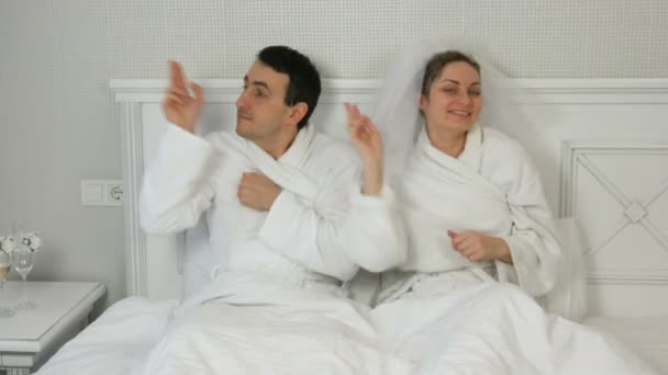 Vicces házaspár friss házasok egy hotelben, akik vidáman táncolnak az ágyban. Menyasszony egy fátyol és egy fehér fürdőköpeny a vőlegény szórakozás és öröm — Stock videók