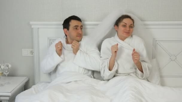 Забавная супружеская пара молодоженов в отеле весело танцуют в постели. Невеста в вуали и белый халат с женихом веселятся и радуются — стоковое видео