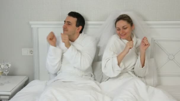 Engraçado casal jovem recém-casados em um hotel alegremente dançando na cama. Noiva em um véu e um roupão de banho branco com o noivo se divertindo e se alegrando — Vídeo de Stock