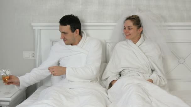 Pareja joven casada divertida de recién casados en el hotel mienten en la cama y beben champán de copas de boda. Novia en un velo y un albornoz blanco con el novio divirtiéndose y regocijándose beso — Vídeos de Stock