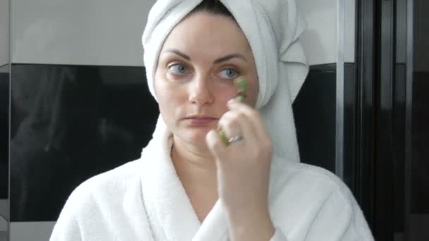 Piękna kobieta z ręcznikiem na głowie robi masaż z jadeitowym wałkiem do twarzy przodem głowy kamienia w łazience. Chińskie narzędzia piękności. Masaż limfatyczny skóry przed starzeniem się zmarszczek — Wideo stockowe